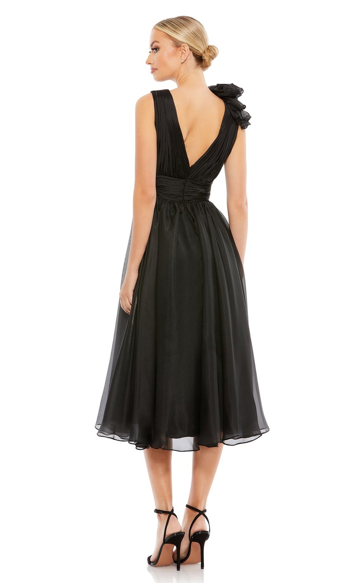 A woman wears the Mac Duggal Short Wedding Guest Dress 70165, a black knee-length dress with a deep V-back, ruched ruffled bodice, and one-shoulder bow detail, paired with black high-heeled sandals as she stands facing away.